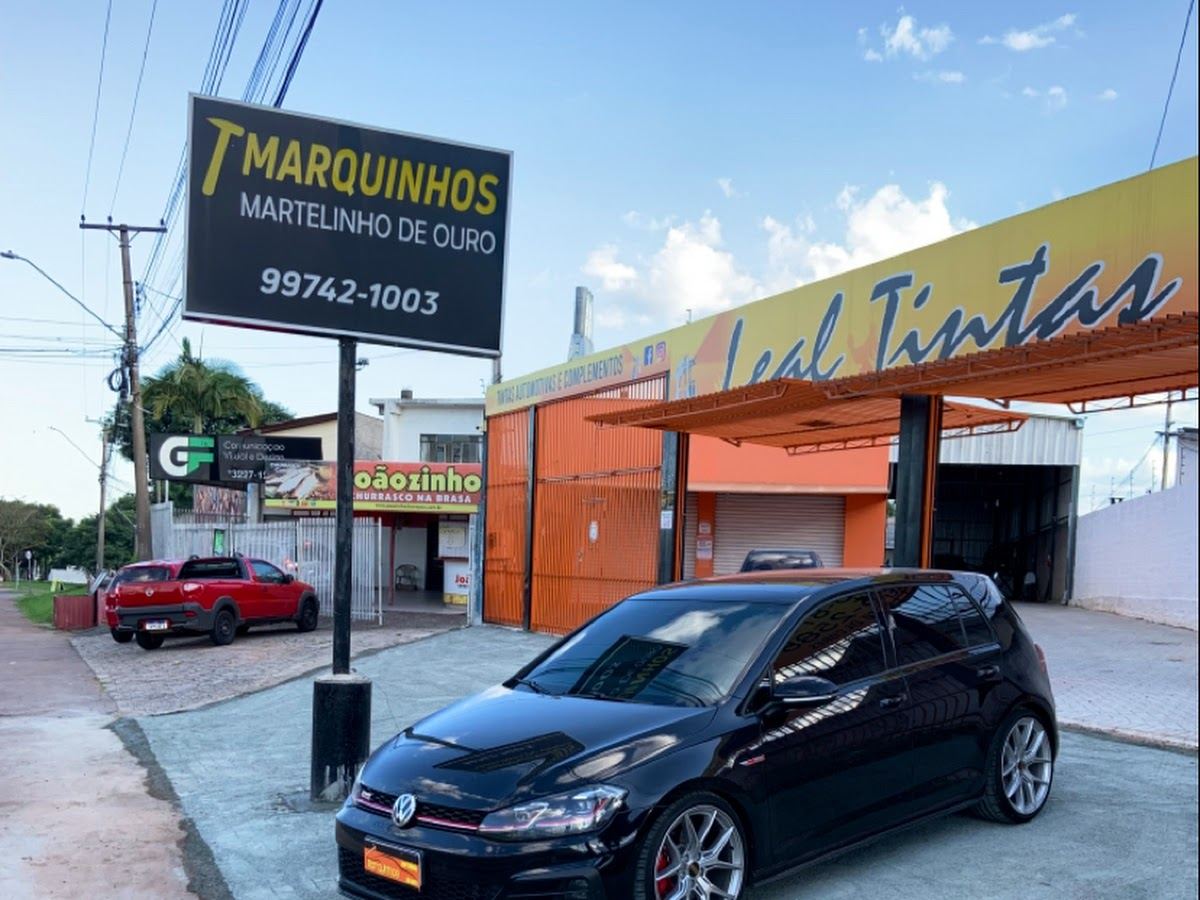 Placa da Marquinhos Martelinho de Ouro na rua, com carros estacionados em frente à oficina